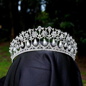 Pode incluir: Uma tiara prateada com cristais transparentes e detalhes de pérolas falsas. A coroa apresenta um design ornamentado com pérolas em forma de gota penduradas na borda inferior. É exibida em uma superfície preta contra um fundo verde desfocado.