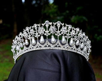 May include: A silver tiara adorned with clear crystals and faux pearls. The crown has an intricate design, with pear-shaped pearls suspended from the lower edge. It is displayed on a black surface, set against a blurred green backdrop.
