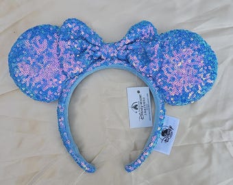 May include: A blue sequined mouse ear headband with a matching bow. The ears and bow are covered in iridescent sequins, creating a shimmering effect. The headband has a tag that says "Disney Resort".