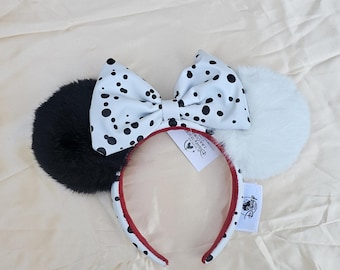 May include: A Dalmatian-themed headband with black and white faux fur ears and a matching bow. The bow and headband are decorated with black spots. The headband has a red trim and a Disney tag.