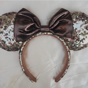 Peut inclure: Un serre-tête avec deux oreilles circulaires recouvertes de paillettes scintillantes dans des tons bronze, cuivre et argent. Un grand nœud central en tissu métallique couleur bronze foncé est placé entre les oreilles. L'arceau est doublé.