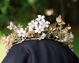 May include: A gold-toned metal crown featuring white floral accents, metallic butterfly embellishments, and small clear crystals. The structure consists of intricate branch-like wirework with scattered pearls and sparkling stones throughout the design.
