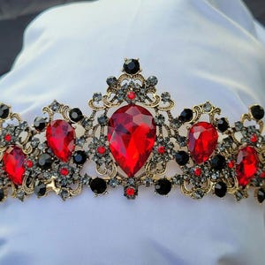 Pode incluir: Uma tiara de metal dourado com um grande cristal vermelho central em forma de pera, rodeado por cristais vermelhos e pretos menores. O intrincado trabalho de filigrana metálica é acentuado com numerosas pequenas pedras cinza-escuras.