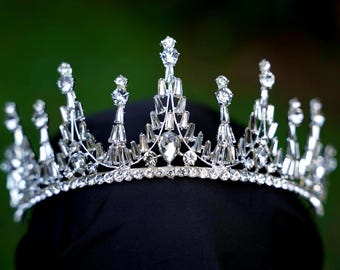 May include: A silver-toned tiara featuring a band encrusted with small clear rhinestones. The crown displays a series of vertical spikes adorned with baguette-cut and round-cut clear crystals, culminating in a central teardrop-shaped stone.