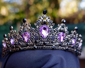 May include: An ornate crown with a black metal framework, embellished with black and purple gemstones. The crown showcases large, teardrop-shaped purple gems and smaller black stones, creating an elegant design. The crown is set against a blurred background.