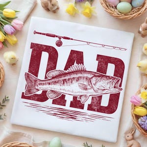 Peut inclure: T-shirt blanc avec un motif bordeaux vieilli. Le motif pr&eacute;sente le mot "DAD" au-dessus d'une illustration d&eacute;taill&eacute;e d'un poisson et d'une canne &agrave; p&ecirc;che. Le t-shirt est entour&eacute; de d&eacute;corations de P&acirc;ques.
