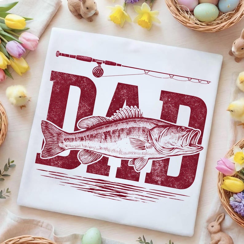 Peut inclure: T-shirt blanc avec un motif bordeaux vieilli. Le motif pr&eacute;sente le mot "DAD" au-dessus d'une illustration d&eacute;taill&eacute;e d'un poisson et d'une canne &agrave; p&ecirc;che. Le t-shirt est entour&eacute; de d&eacute;corations de P&acirc;ques.