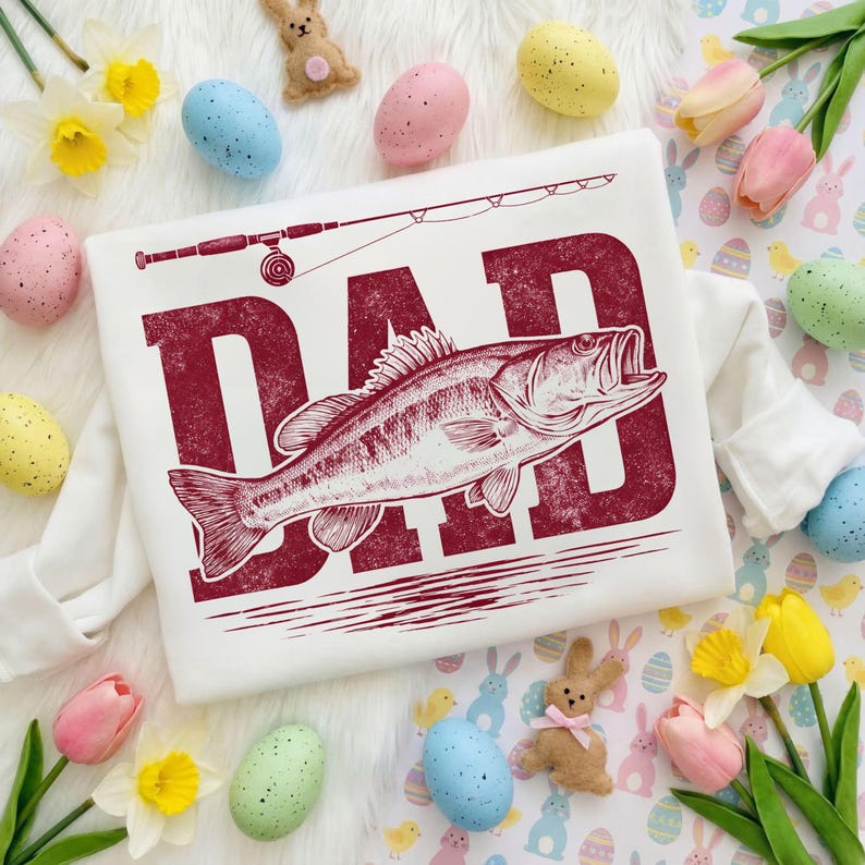 Peut inclure: T-shirt blanc &agrave; manches longues avec un motif bordeaux vieilli indiquant "DAD", une illustration de poisson et une canne &agrave; p&ecirc;che. Le t-shirt est entour&eacute; d'&oelig;ufs de P&acirc;ques, de fleurs et de d&eacute;corations de lapins.