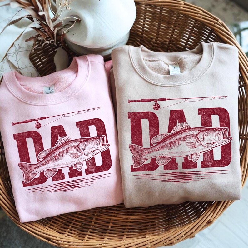 Peut inclure: Deux sweat-shirts &agrave; col rond, un rose clair et un beige, avec un motif rouge. Le motif repr&eacute;sente le mot "DAD" au-dessus d'un poisson et d'une canne &agrave; p&ecirc;che. Les sweat-shirts sont pr&eacute;sent&eacute;s dans un panier en osier.