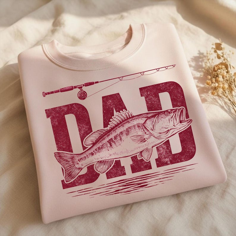 Peut inclure: Un sweat-shirt rose clair avec un motif bordeaux vieilli. Le motif repr&eacute;sente le mot "DAD" avec un poisson et une canne &agrave; p&ecirc;che. Le sweat-shirt est fait d'une mati&egrave;re douce.