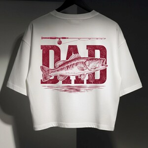 Peut inclure: T-shirt blanc avec un motif graphique rouge repr&eacute;sentant le mot "DAD", un poisson et une canne &agrave; p&ecirc;che. Le motif est centr&eacute; au dos du t-shirt. Le t-shirt est suspendu &agrave; un cintre noir.