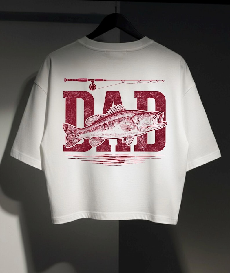 Peut inclure: T-shirt blanc avec un motif graphique rouge repr&eacute;sentant le mot "DAD", un poisson et une canne &agrave; p&ecirc;che. Le motif est centr&eacute; au dos du t-shirt. Le t-shirt est suspendu &agrave; un cintre noir.