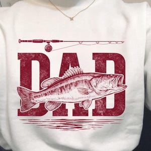 Peut inclure: Sweat-shirt blanc avec un motif rouge repr&eacute;sentant un poisson, une canne &agrave; p&ecirc;che et le mot "DAD". Le poisson est d&eacute;taill&eacute; avec des &eacute;cailles et des nageoires. La canne &agrave; p&ecirc;che est au-dessus du mot "DAD".