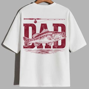 Peut inclure: T-shirt blanc avec un motif rouge repr&eacute;sentant le mot "DAD", un poisson et une canne &agrave; p&ecirc;che. Le motif est centr&eacute; au dos du t-shirt. Le t-shirt a des manches courtes et un col rond.