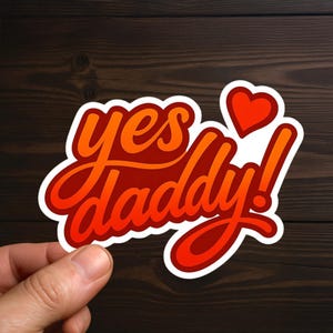 May include: A sticker with the words "yes daddy!" in a stylized, cursive font, with a red and orange gradient. A small red heart is above the exclamation point. The sticker has a white border.