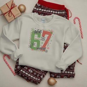 May include: A cream-coloured sweatshirt featuring the text "six 67 seven" in green and red, adorned with snowflakes and lights. It's styled with red and white patterned pyjama bottoms, a wrapped gift, and Christmas decorations.