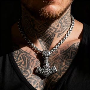 Op de afbeelding: Een zilverkleurige ketting met een hanger in de vorm van Thors hamer, Mjolnir. De hanger is gedetailleerd met Viking-ontwerpen en symbolen. De ketting heeft een dikke, gevlochten stijl. De ketting wordt tegen een donkere achtergrond gedragen.