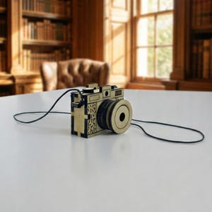 May include: A wooden camera with a black cord, set on a white table. The camera has a gold-colored lens and body, with intricate cut-out details. The background features a library with bookshelves and a window.