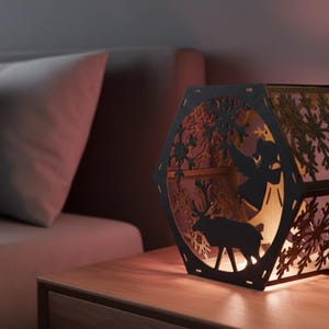 May include: A black, hexagonal, laser-cut wooden lantern with a winter scene featuring an angel and a reindeer. The lantern is illuminated from within, casting a warm glow. The design includes snowflake cutouts.
