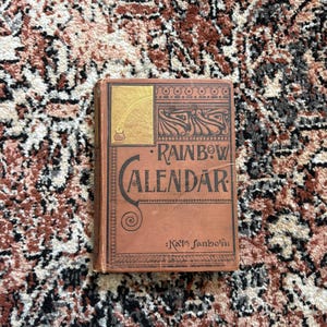 May include: A vintage "Rainbow Calendar" book with a brown cover and gold accents. The title is in black lettering with decorative swirls. The author's name, Kate Sanborn, is printed below the title. The book is on a patterned rug.