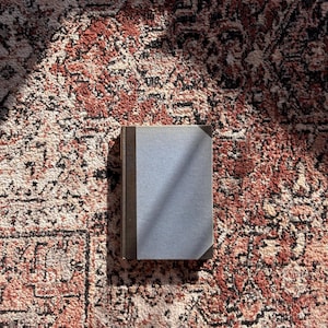 May include: A closed book with a light blue cover and a dark brown spine and edges. The book is lying on a patterned rug with shades of red, brown, and cream.