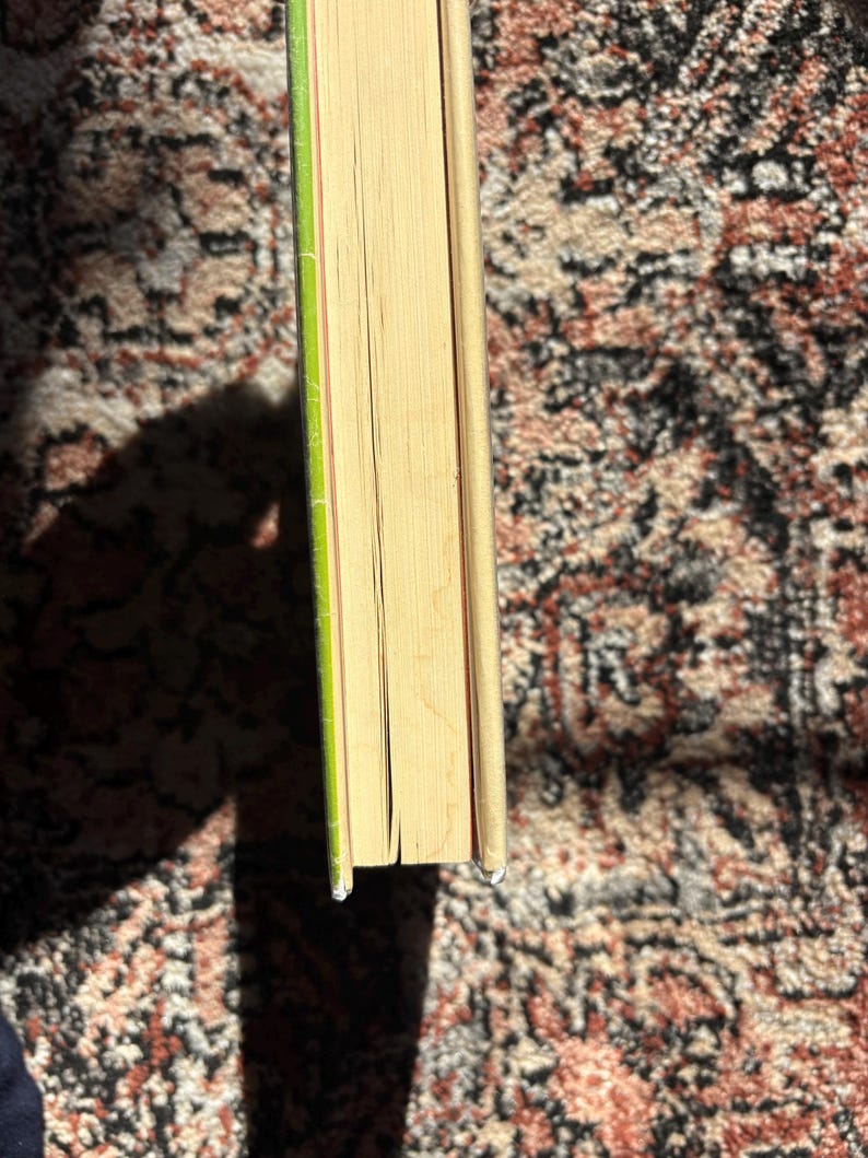 Op de afbeelding: Een hardcover boek met vergeelde pagina's en een groene rug. De randen van het boek zijn zichtbaar en tonen de dikte van de pagina's. Het boek ligt op een patroonkleed, met een schaduw.