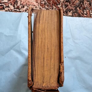 May include: A vintage book with a worn, brown leather cover and aged pages. The spine is partially detached, revealing the book's interior. The edges of the pages are yellowed, indicating age. The book is resting on a light blue surface.