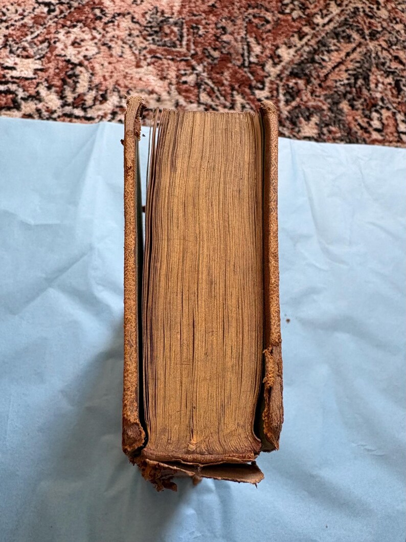 May include: A vintage book with a worn, brown leather cover and aged pages. The spine is partially detached, revealing the book's interior. The edges of the pages are yellowed, indicating age. The book is resting on a light blue surface.