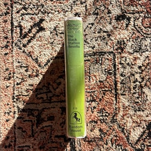 Op de afbeelding: Een hardcover boek met een groene rug, getiteld "The Black Stallion Revolts" van Walter Farley. Het boek heeft witte tekst en een Random House-logo met een paard. Het boek ligt op een patroonkleed.
