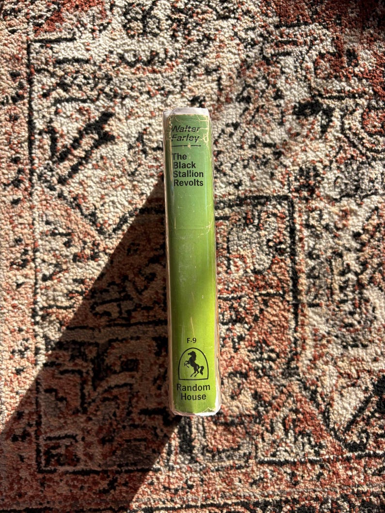 Op de afbeelding: Een hardcover boek met een groene rug, getiteld "The Black Stallion Revolts" van Walter Farley. Het boek heeft witte tekst en een Random House-logo met een paard. Het boek ligt op een patroonkleed.