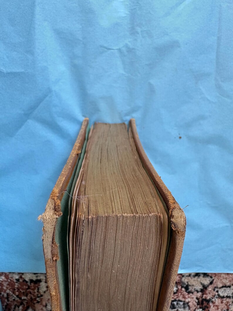 May include: A vintage book with aged, yellowed pages and a worn, brown leather cover. The edges of the cover show signs of wear and tear, with the spine slightly damaged. The book's pages are tightly bound, suggesting it is a hardcover edition.