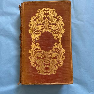 May include: Antique book with a brown cover and gold filigree design. The cover features a symmetrical, ornate floral pattern. The book's edges show signs of wear, suggesting its age. The background is a light blue surface.