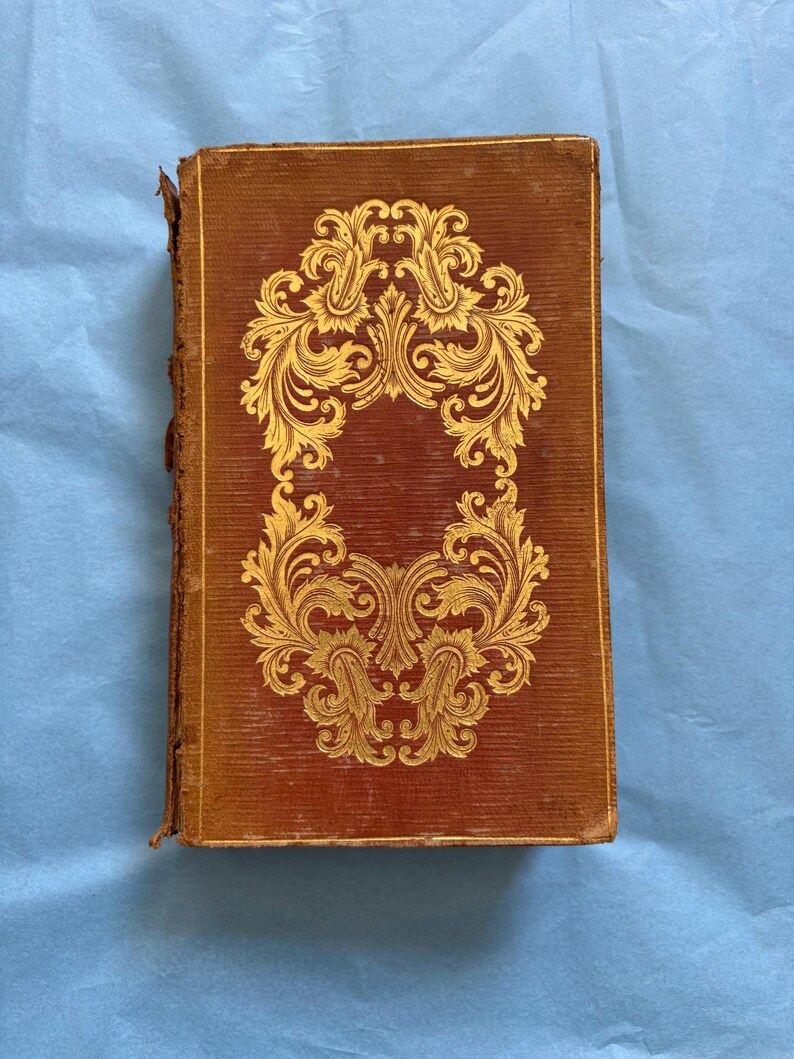 May include: Antique book with a brown cover and gold filigree design. The cover features a symmetrical, ornate floral pattern. The book's edges show signs of wear, suggesting its age. The background is a light blue surface.