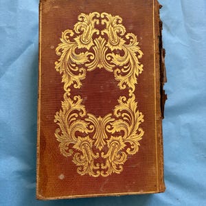 May include: Antique book with a worn, brown leather cover. The cover features a gold, ornate floral design in the center. The edges of the book show signs of wear and age, adding to its vintage appeal. The book is rectangular in shape.
