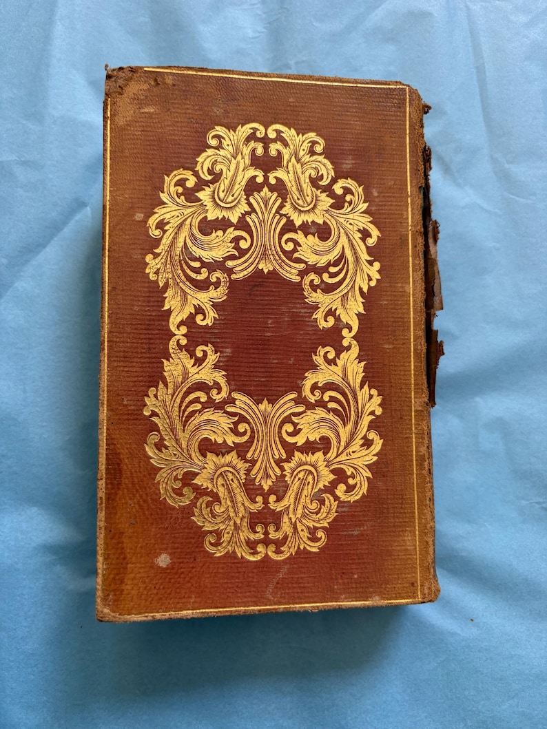 May include: Antique book with a worn, brown leather cover. The cover features a gold, ornate floral design in the center. The edges of the book show signs of wear and age, adding to its vintage appeal. The book is rectangular in shape.