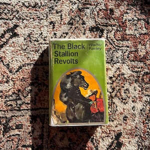 Op de afbeelding: Een vintage hardcover boek getiteld "The Black Stallion Revolts" van Walter Farley. De omslag toont een zwart paard en een cowboy in een rood overhemd tegen een oranje achtergrond, in een groen kader.