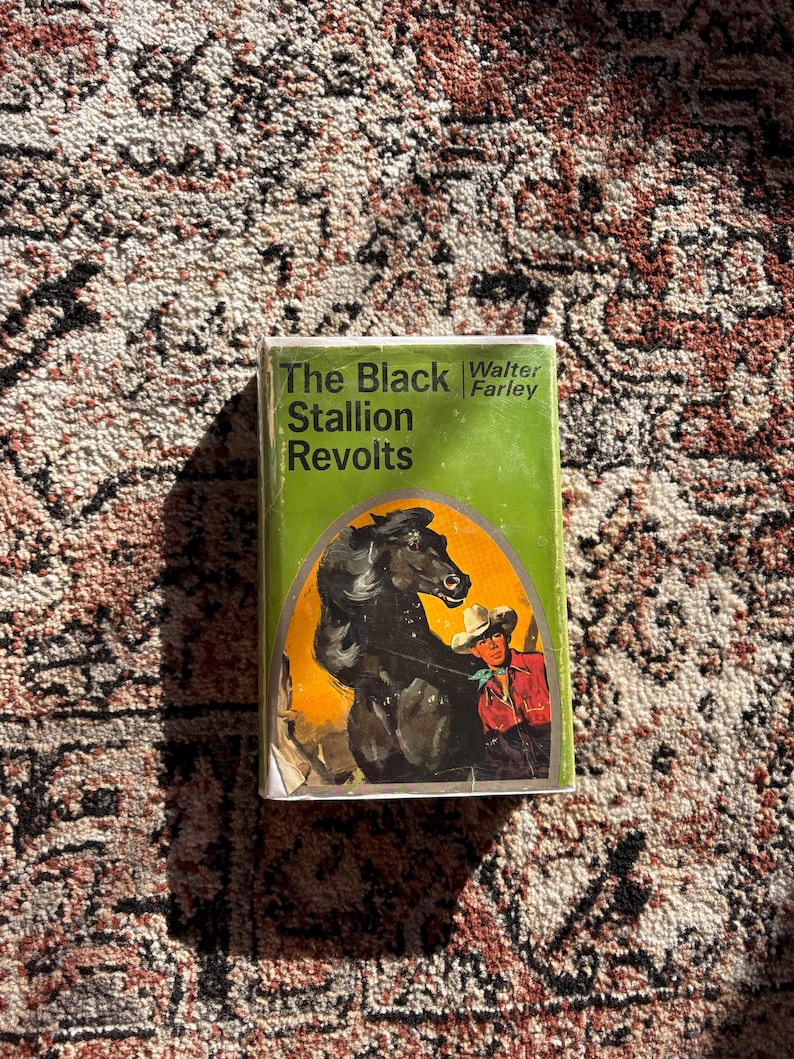 Op de afbeelding: Een vintage hardcover boek getiteld "The Black Stallion Revolts" van Walter Farley. De omslag toont een zwart paard en een cowboy in een rood overhemd tegen een oranje achtergrond, in een groen kader.