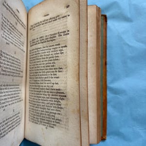 May include: An open antique book with aged, yellowed pages. The book's spine is visible, showing a brown leather cover with green accents. Text is printed on the pages, and the book is angled, revealing the edges of the pages.