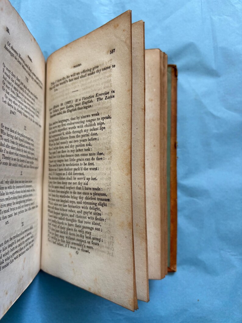 May include: An open antique book with aged, yellowed pages. The book's spine is visible, showing a brown leather cover with green accents. Text is printed on the pages, and the book is angled, revealing the edges of the pages.