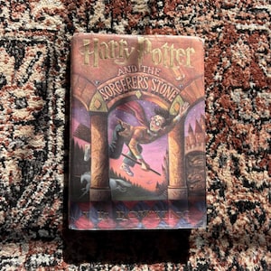 May include: A hardcover book titled "Harry Potter and the Sorcerer's Stone" with a colorful illustration of Harry Potter flying on a broomstick. The cover features a dark red and gold color scheme, with the title prominently displayed.