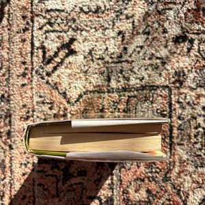 Op de afbeelding: Een hardcover boek met een witte stofomslag, liggend op een patroonkleed. De pagina's van het boek zijn vergeeld en de rug is zichtbaar. Het kleed heeft een bruin, rood en beige ontwerp.