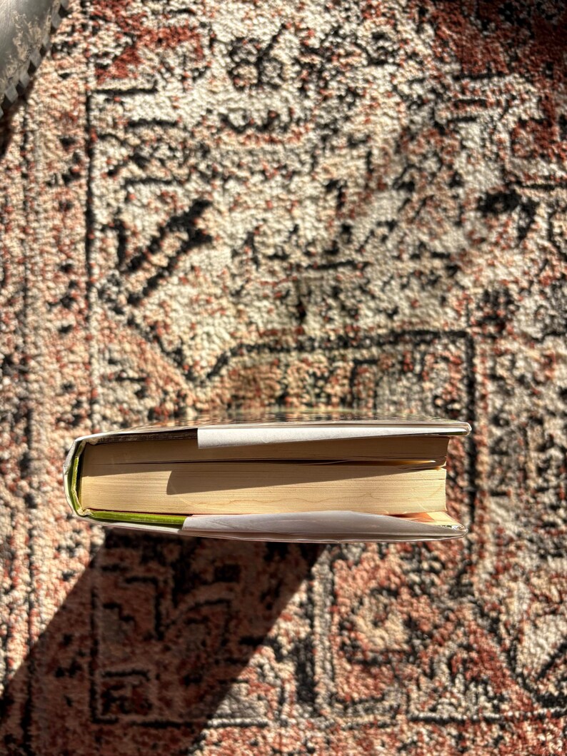 Op de afbeelding: Een hardcover boek met een witte stofomslag, liggend op een patroonkleed. De pagina's van het boek zijn vergeeld en de rug is zichtbaar. Het kleed heeft een bruin, rood en beige ontwerp.