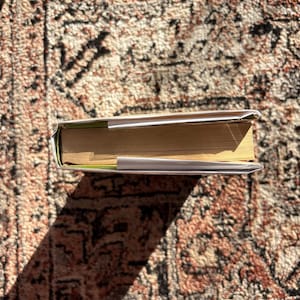 Op de afbeelding: Een gesloten boek met een witte kaft en vergeelde pagina's, liggend op een patroonkleed. De rug van het boek is zichtbaar en de pagina's zijn iets uitgespreid. Het kleed heeft een rood en bruin kleurenschema.