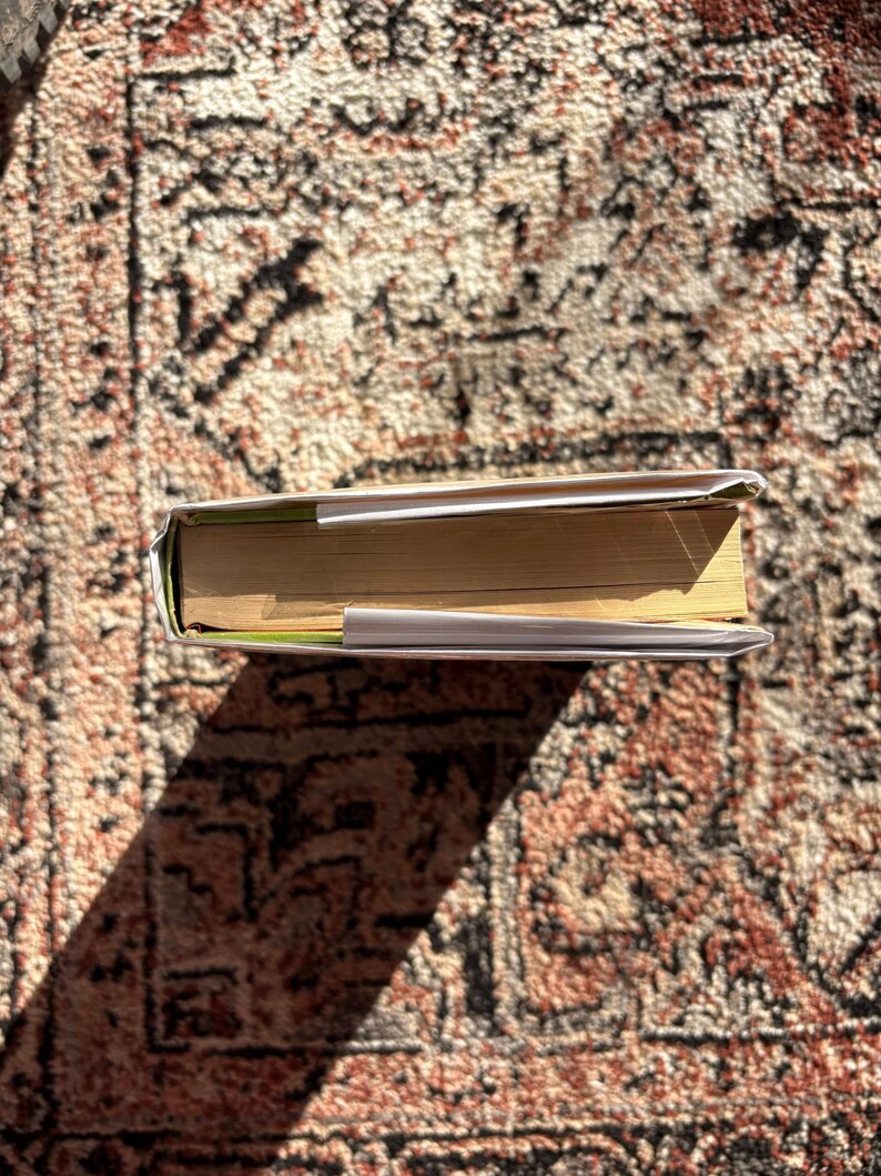 Op de afbeelding: Een gesloten boek met een witte kaft en vergeelde pagina's, liggend op een patroonkleed. De rug van het boek is zichtbaar en de pagina's zijn iets uitgespreid. Het kleed heeft een rood en bruin kleurenschema.