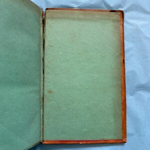 May include: An open antique book with a teal-colored interior and a brown, worn cover. The pages are aged, showing signs of wear and use. The book's spine is visible, revealing its binding.