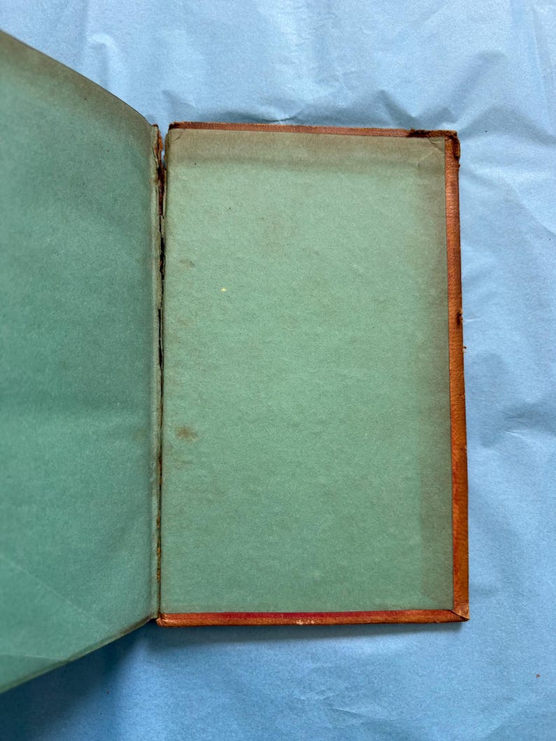 May include: An open antique book with a teal-colored interior and a brown, worn cover. The pages are aged, showing signs of wear and use. The book's spine is visible, revealing its binding.