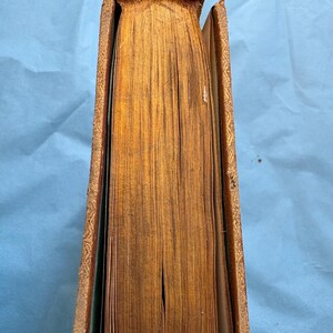 May include: A vintage book with aged, yellowed pages and a worn, brown leather cover. The spine is damaged, and the edges of the pages are gilded. The cover has gold embossed detailing.