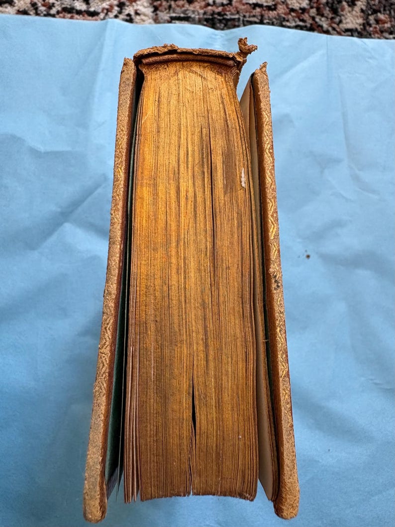 May include: A vintage book with aged, yellowed pages and a worn, brown leather cover. The spine is damaged, and the edges of the pages are gilded. The cover has gold embossed detailing.