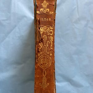 May include: A vintage book spine with the title "ILTON" in gold lettering. The cover is brown leather with ornate gold floral designs. The book shows signs of age and wear, with some damage to the edges.