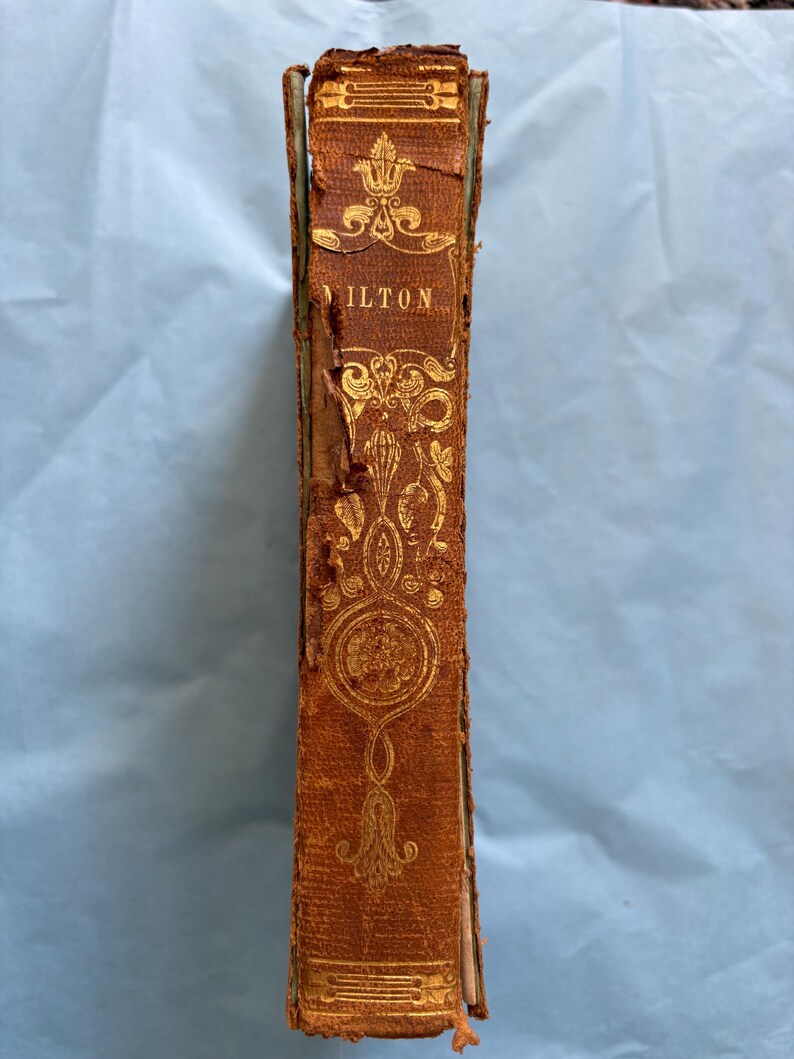 May include: A vintage book spine with the title "ILTON" in gold lettering. The cover is brown leather with ornate gold floral designs. The book shows signs of age and wear, with some damage to the edges.